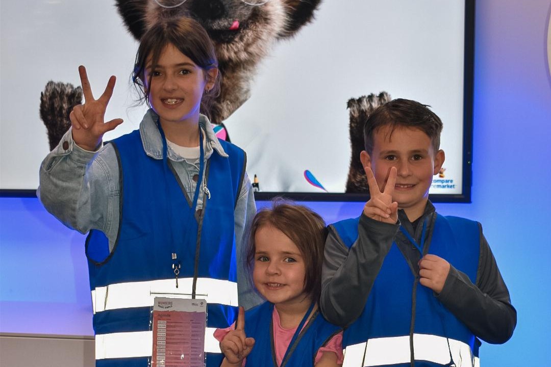 Three children smile while standing in front of a Compare the Market image of meerkat Sergei