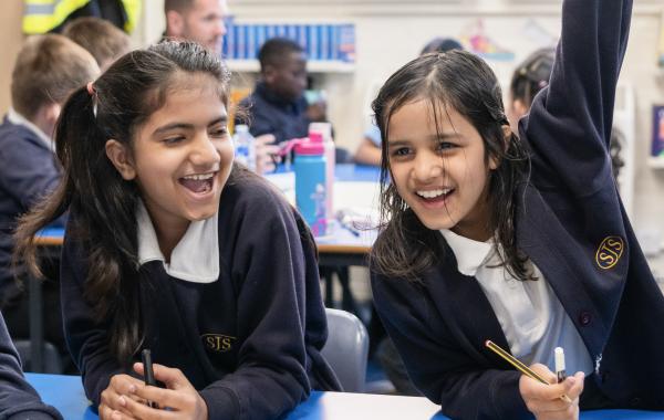 Two school pupils sitting at a table looking happy, one has their arm raised as if to answer a question