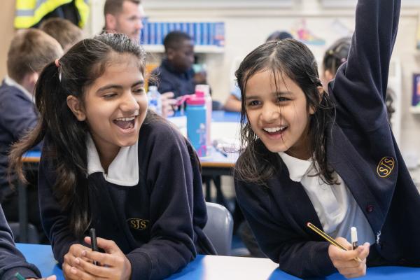 Two school pupils sitting at a table looking happy, one has their arm raised as if to answer a question